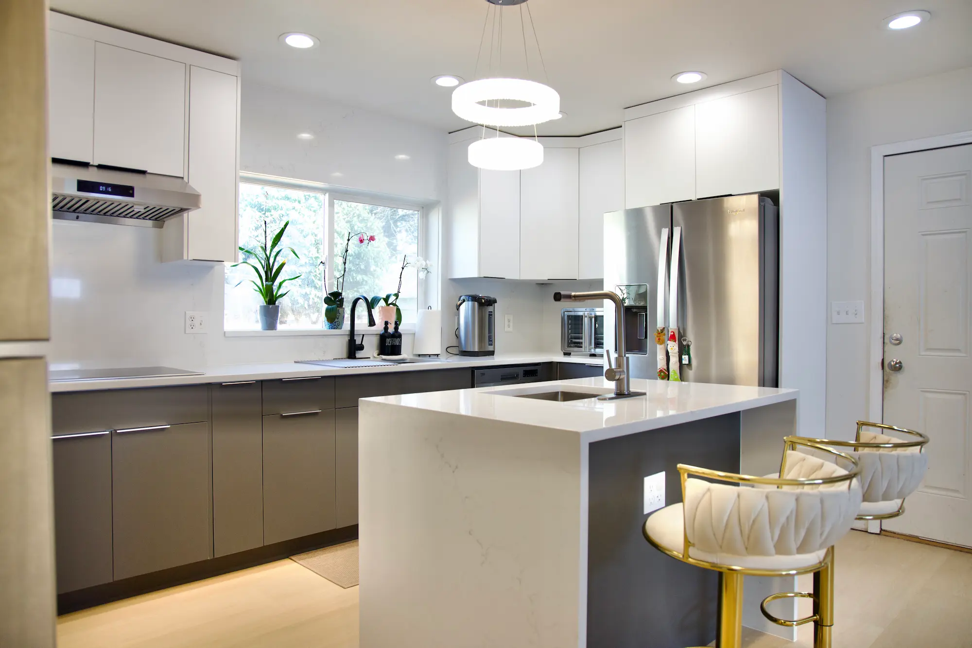Modern kitchen with white upper cabinets, gray lower cabinets, stainless steel fridge, range hood, island with sink, and white stools with gold bases.