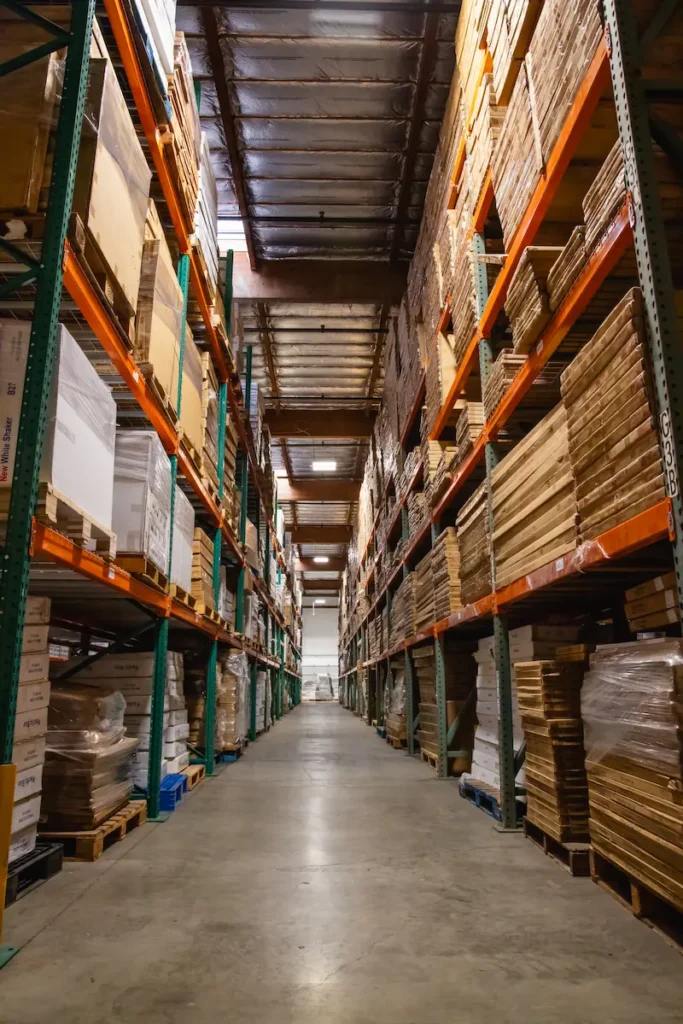 Warehouse aisle with tall shelves stacked with wooden boards and boxes, concrete floor, and overhead lighting.